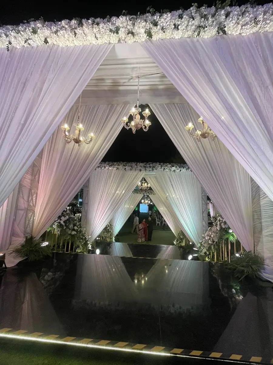 A symmetrical mandap corridor draped in white fabric with crystal chandeliers hanging under each peak, the floor a black mirrored surface reflecting the lights, lit for the evening.