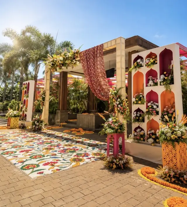 A daytime mandap with a wall of arched floral niches in maroon, orange and white, a patterned rug on the ground and palm trees behind.