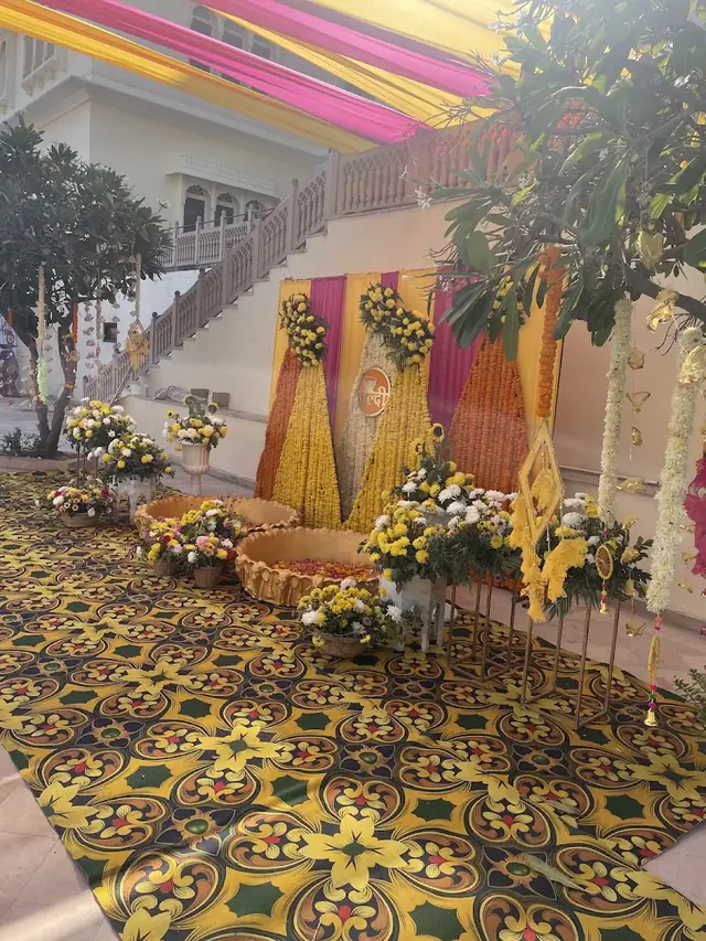 A haldi-style backdrop of three marigold and pink floral panels with woven cane baskets of loose petals on a patterned yellow rug.