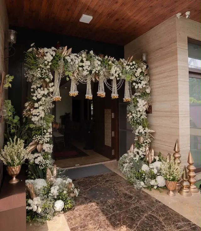 A doorway framed by a white floral arch with hanging bead and gold tassel strands, brass urns of white flowers on either side.