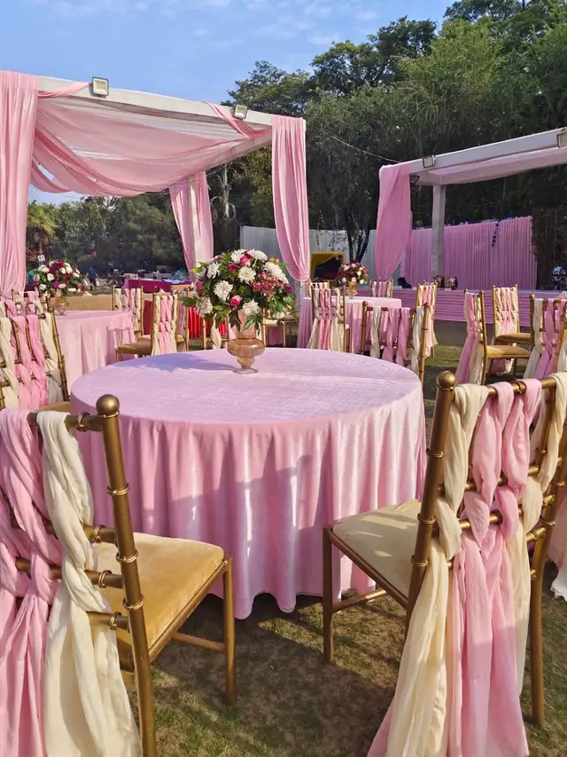 Round tables in a pink theme with draped pink and cream chair covers, under a pink fabric canopy outdoors.