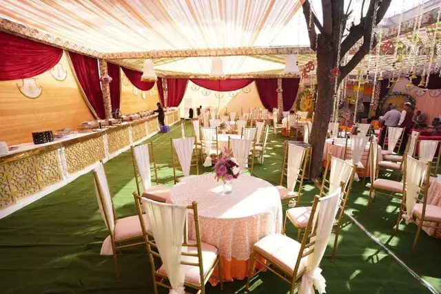 A night-time stage with band equipment and a floral arrangement beneath a tree strung with chandeliers and marigold strands.